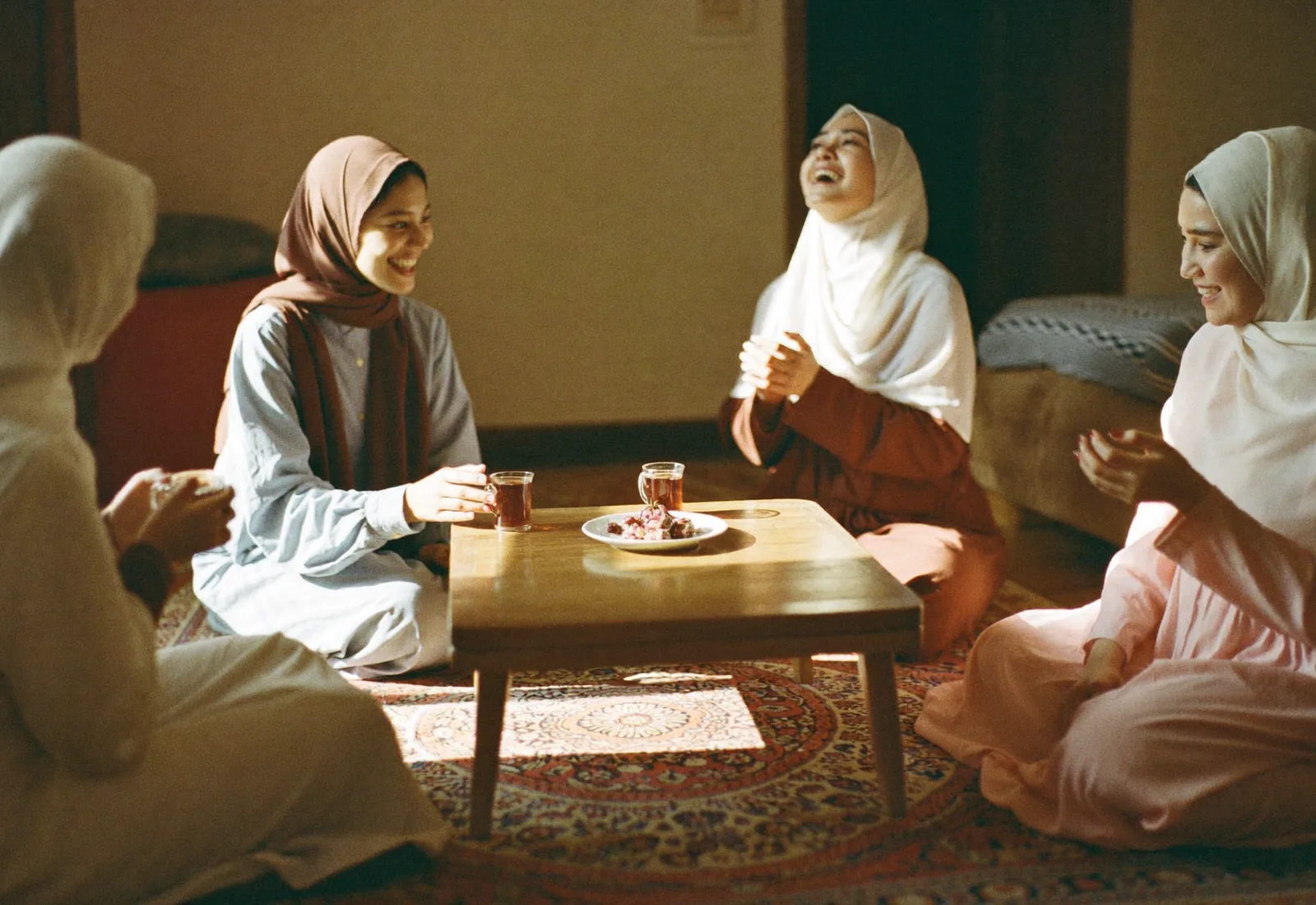 Four women in hijab sitting on a patterned rug around a low table, laughing together over glasses of tea