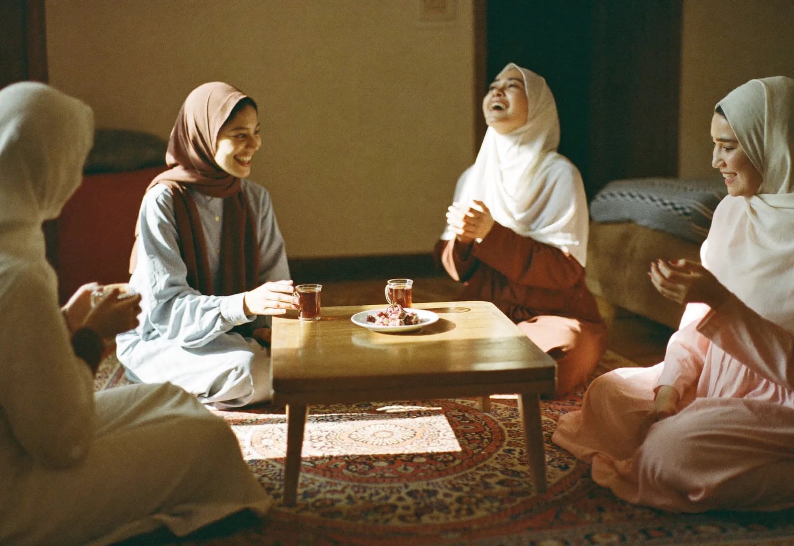 Four women in hijab sitting on a patterned rug around a low table, laughing together over glasses of tea