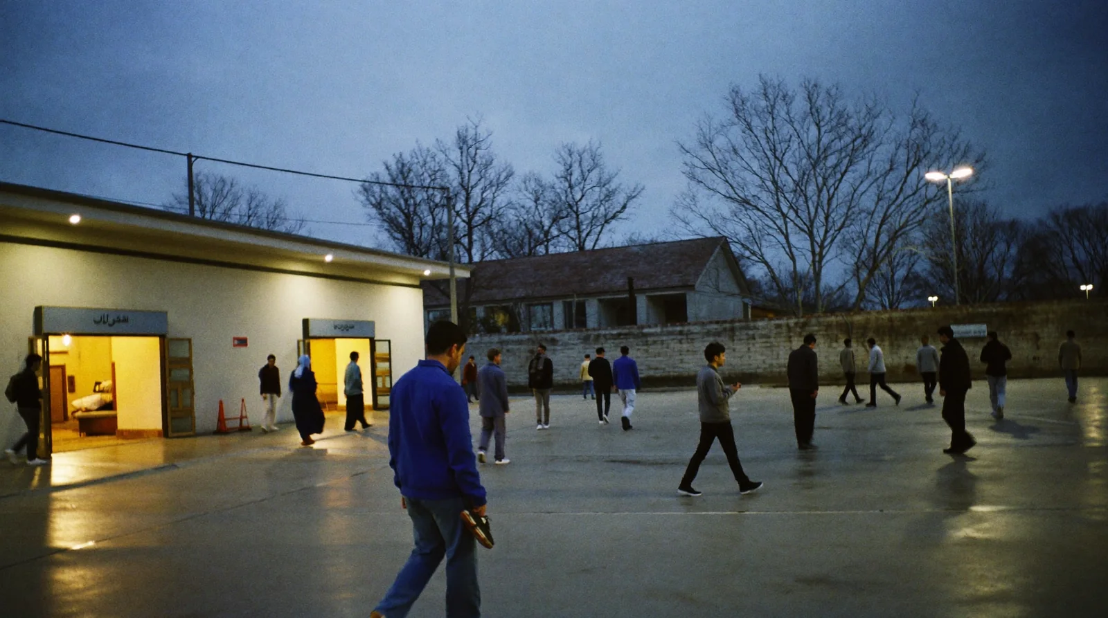 Men leaving a masjid courtyard in ones and twos after prayer at dusk, shoes in hand, nobody stopping to talk